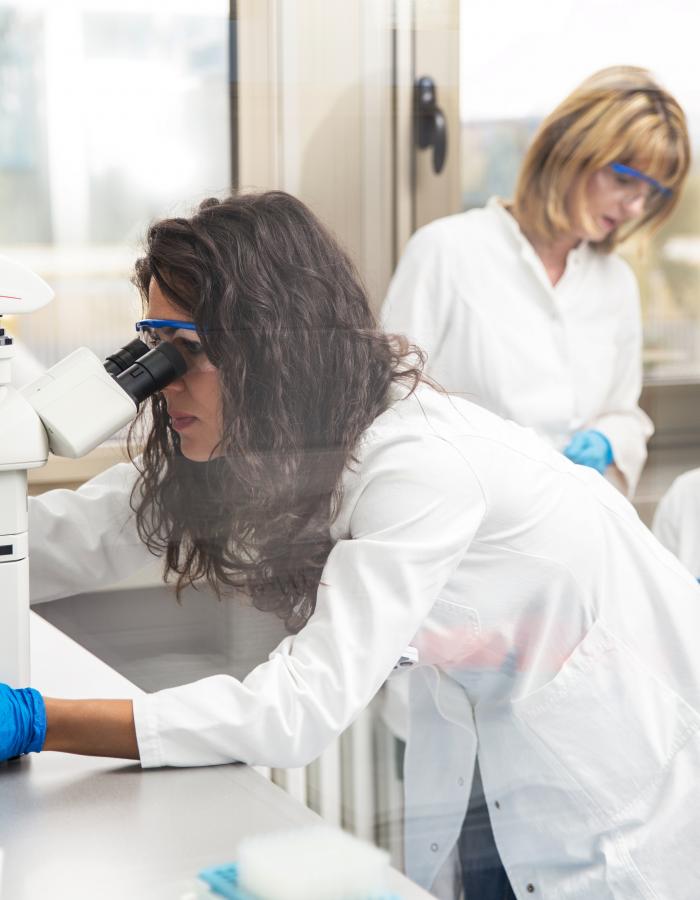 A photo of a female scientist looking through a microscope with two female scientists in the background.