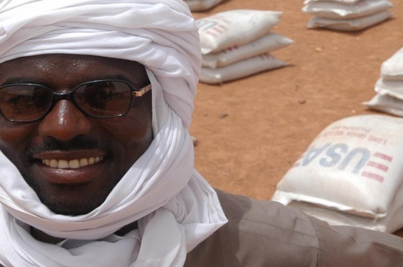 A photo of a humanitarian worker smiling with packs of food aid in the background.